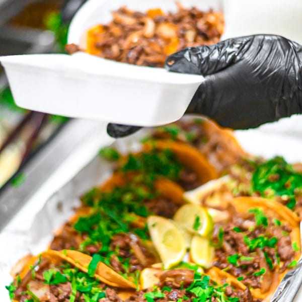 Gloved hand serving a white takeout container of Mexican food, with tacos in the foreground.