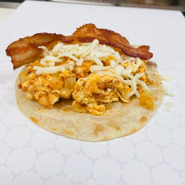 A breakfast taco with scrambled eggs, potatoes, shredded cheese, and a strip of bacon.