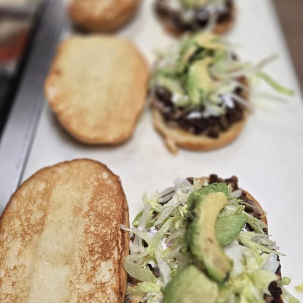 Several Mexican tortas with meat, lettuce, avocado, and crema, with toasted buns beside them.